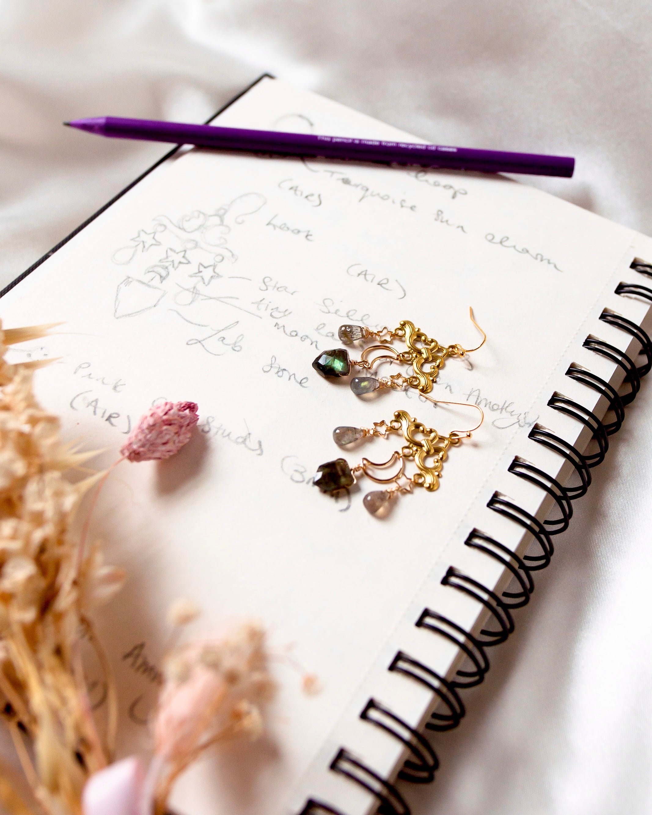 Personalised custom jewellery for gifts. Image shows design process of Wanderlust earrings sketches with the earrings placed on top. The earrings feature golden filigree clouds, Labradorite, stars and crescent moons and hooks. A pencil is placed next to the design sketches in the notepad and dried flowers sit atop the paper.