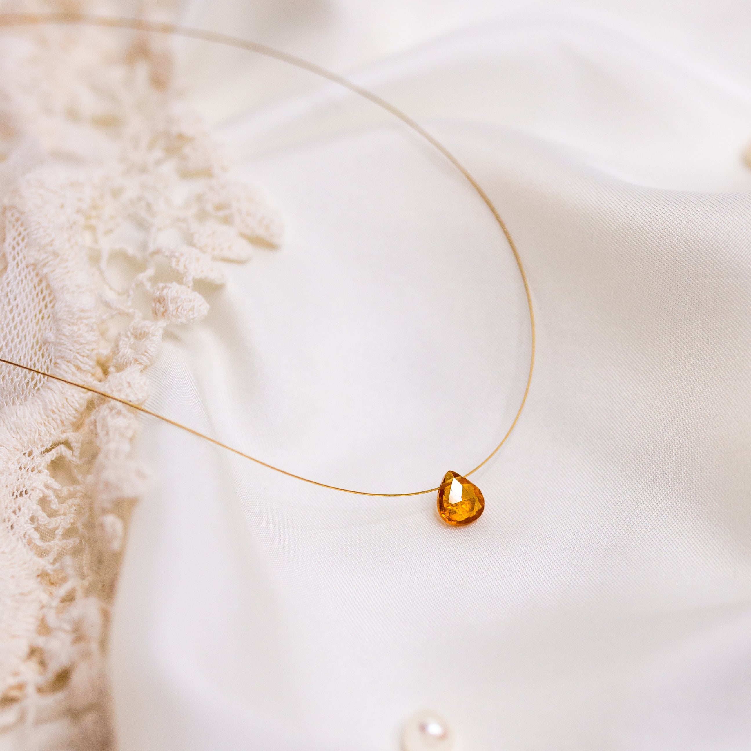 A gold coloured floating crystal necklace with a orange garnet coloured pendant, displayed delicately over a white fabric surface.